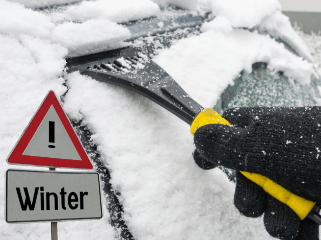 Iemand maakt de ruit van zijn auto sneeuwvrij, ook zie je een verkeersbord met de tekst 'Winter'.